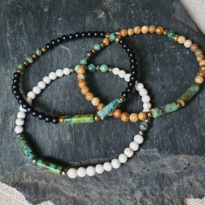 May include: Three bracelets made of different beads. One bracelet is black, one is white, and one is brown with green and gold accents.