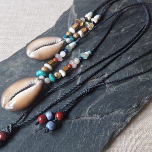 Può includere: Due collane con ciondoli a conchiglia di cauri e accenti di perline colorate. Le collane presentano un cordino nero e una varietà di perline nelle tonalità del blu, turchese, bianco e marrone. Le conchiglie di cauri sono di colore beige chiaro.