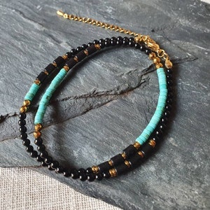 Könnte beinhalten: Eine mehrreihige Halskette mit schwarzen Perlen, türkisfarbenen zylindrischen Perlen und goldfarbenen Akzenten. Die Halskette hat eine zarte goldene Kette und einen Verschluss. Der Schmuck wird auf einer dunkelgrauen Oberfläche präsentiert.