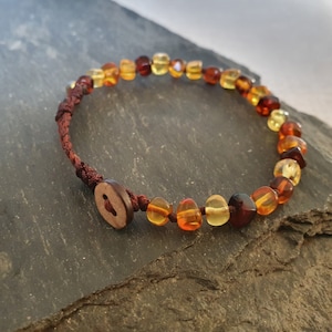 Puede incluir: Un brazalete de cordón marrón con cierre de botón de madera y cuentas de ámbar en tonos de amarillo, naranja y rojo.