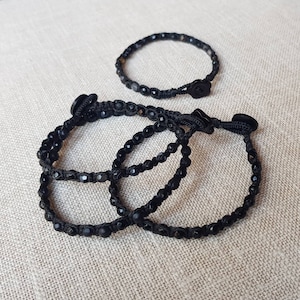 Op de afbeelding: Drie zwarte kralen armbanden met zwart koord en een zwarte knoopsluiting. De armbanden zijn op elkaar gelegd.