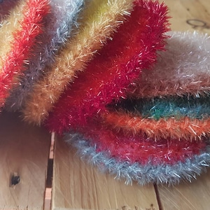 Puede incluir: Una pila de paños de cocina coloridos y brillantes hechos de hilo de crochet. Los paños de cocina son en tonos de azul, morado, rojo, naranja, amarillo y verde.