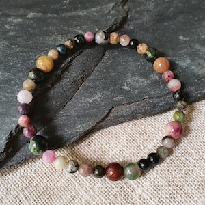 Puede incluir: Un brazalete de piedras preciosas multicolores con cuentas de varios tamaños y colores, incluyendo rosa, verde, marrón, negro y dorado. El brazalete está hecho de piedras naturales y está enhebrado en un cordón elástico transparente.