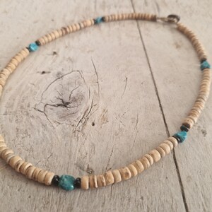 Puede incluir: Un collar que presenta una serie de pequeñas cuentas de color tostado claro en forma de disco, intercaladas con piedras de color turquesa brillante y pequeñas cuentas negras redondas. El collar se asegura con un cierre metálico sencillo.