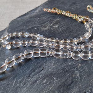 Peut inclure: Collier de perles en cristal transparent avec un pompon décoratif. Le collier présente une variété de perles transparentes, rondes et rectangulaires, enfilées ensemble. Les perles sont reliées par une fine ficelle dorée. Le pompon est fait d'un cordon torsadé multicolore.