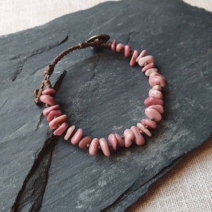 Puede incluir: Un brazalete de piedra rosa y blanca con un cordón de cuero marrón y un cierre de metal. El brazalete está sobre una superficie de pizarra gris oscuro.