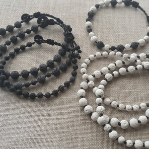 Könnte beinhalten: Zwei Lava-Perlenarmbänder, eines schwarz und eines weiß. Die Armbänder bestehen aus kleinen, runden Perlen und sind mit schwarzem Band zusammengebunden.