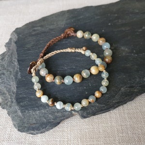 Puede incluir: Dos pulseras hechas con cordón marrón y beige y cuentas de piedras preciosas azul claro y marrón.