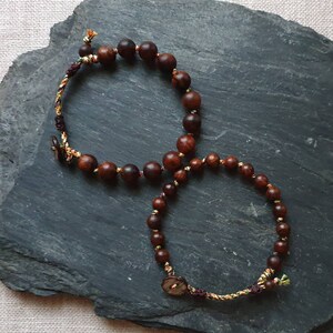 May include: Two brown wooden bead bracelets with colorful string accents. The bracelets are tied together with a knot and have a small button on each end.