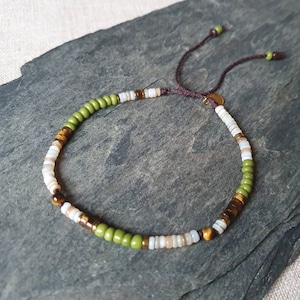 Könnte beinhalten: Ein Armband mit Perlen aus einer Mischung aus grünen, braunen, weißen und goldenen Perlen. Das Armband hat einen Schiebeknotenverschluss mit braunen Kordeln und kleinen grünen Perlen an den Enden. Die Perlen sind auf eine flexible Schnur gefädelt.