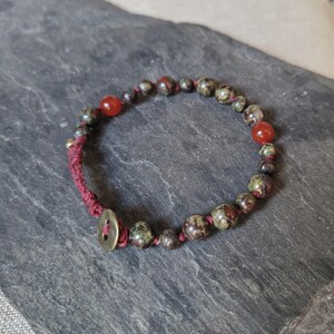 Peut inclure: Un bracelet de perles composé de perles vert foncé et rouge-brun, entrecoupées de perles rouges vives. Le bracelet est monté sur un cordon rouge foncé et fermé par un fermoir bouton de couleur laiton. Circonférence d'environ 18 cm.