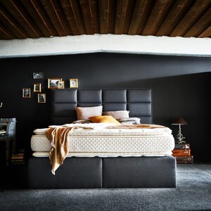 Könnte beinhalten: Ein modernes Bett mit einem dunkelgrauen, gepolsterten Kopfteil und Sockel. Das Bett verfügt über eine weiße, gesteppte Matratze und ist mit Kissen und einer beigen Decke geschmückt. Der Raum hat eine dunkelgraue Wand und eine Holzdecke.