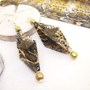 Könnte beinhalten: Ein Paar geometrische Ohrhänger mit Schlangenmuster in Schwarz, Braun und Gold. Die Ohrringe haben eine rautenförmige Form mit einem kleinen quadratischen Akzent und einer goldenen Perle unten. Die Ohrhaken sind silberfarben.