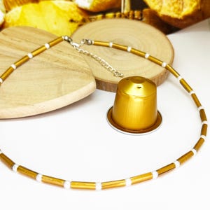 Könnte beinhalten: Eine goldfarbene Halskette mit weißen Akzenten, präsentiert mit einer goldenen Kaffeekapsel. Die Halskette besteht aus kleinen, rechteckigen Perlen und weißen zylindrischen Perlen. Die Halskette liegt auf einer weißen Oberfläche mit Holzakzenten.
