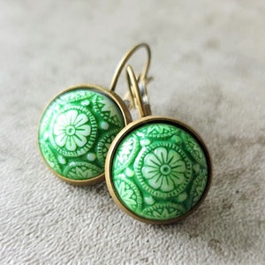 Puede incluir: Un par de pendientes redondos con un diseño floral verde. Los pendientes tienen un marco de metal color latón y un cierre de palanca. El diseño verde presenta una flor central y patrones circundantes.
