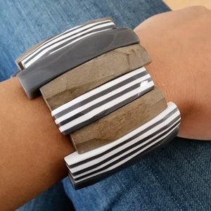 Puede incluir: Un brazalete grueso con segmentos rectangulares alternos. El brazalete presenta segmentos en tonos grises y marrones, con detalles a rayas blancas y negras. El brazalete se lleva en una muñeca, con unos vaqueros en el fondo.