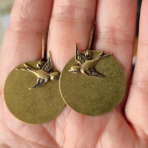 Puede incluir: Un par de pendientes de color latón. Cada pendiente presenta un colgante circular con una silueta detallada de un pájaro en vuelo. Los pájaros están unidos a un disco circular. Los pendientes tienen un cierre de gancho.