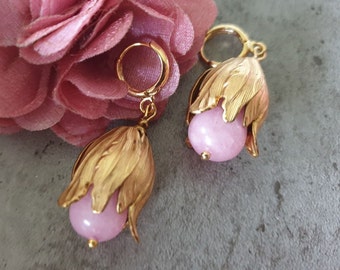 May include: A pair of gold-colored earrings featuring a flower-like design. Each earring has a light pink, round bead at the center, surrounded by gold-colored petals. The earrings are on a gray surface, with a pink flower in the background.