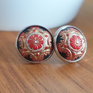 Puede incluir: Un par de pendientes de botón redondos con borde plateado. Los pendientes presentan un diseño detallado con detalles en rojo, dorado y negro. El diseño central es una flor roja con un centro blanco.
