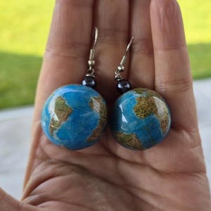 Könnte beinhalten: Ein Paar Hängeohrringe mit einem blauen und braunen Globus-Design. Die Globen sind an silbernen Haken mit einer schwarzen Perle an der Oberseite jedes Hakens befestigt.