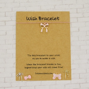 May include: A tan card with the words "Wish Bracelet" printed at the top. A pink string is strung across the card with a bow charm. The text on the card reads, "Tie this bracelet to your wrist, as you do make a wish."