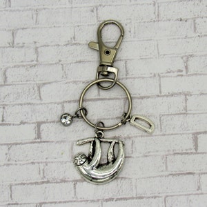 May include: Silver sloth keychain with a silver ring, a silver charm, and a clear rhinestone charm. The keychain is hanging from a silver lobster clasp.