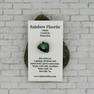 May include: A small green and black rainbow fluorite crystal on a white card with the text "Rainbow Fluorite", "Clarity", "Creativity", "Protection", "Also Aides in: Calming, intuition and mind-body-spirit connection. Works well with: Amethyst", "Water Safe: No", "Sun Safe: No", and "www.salemwishes.com".