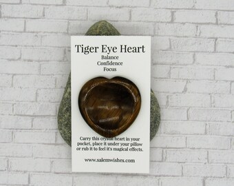 May include: Tiger Eye Heart crystal, carved in the shape of a heart, on a white card with the text "Tiger Eye Heart, Balance, Confidence, Focus, Carry this crystal heart in your pocket, place it under your pillow or rub it to feel it's magical effects. www.salemwishes.com".