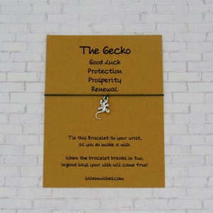 May include: A brown card with the text "The Gecko" and a list of words: "Good Luck", "Protection", "Prosperity", "Renewal". A green string bracelet with a silver gecko charm is attached to the card. The card also includes the text: "Tie this Bracelet to your wrist, as you do make a wish. When the bracelet breaks in two, legend says your wish will come true! salemwishes.com"
