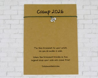May include: A dark green string bracelet featuring a small silver-toned charm shaped like a signpost with the word CAMP on it. The bracelet is attached to a rectangular tan card printed with the text Camp 2026 and a wish-related poem.