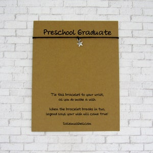 May include: A brown card with the text "Preschool Graduate" printed on it. A black string bracelet with a silver star charm is attached to the card. The text on the card reads: "Tie this bracelet to your wrist, as you do make a wish. When the bracelet breaks in two, legend says your wish will come true! Salemwishes.com"