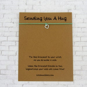 May include: A brown card with the text "Sending You A Hug" printed on it. The card has a light blue string with a silver heart charm attached to it. The text on the card reads: "Tie this bracelet to your wrist, as you do make a wish. When the bracelet breaks in two, legend says your wish will come true! salemwishes.com"