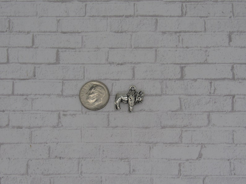 May include: A silver-toned metal charm shaped like a bison, featuring a small loop at the top for attachment. The charm is positioned next to a United States dime, which measures approximately 0.705 inches in diameter, for scale reference.