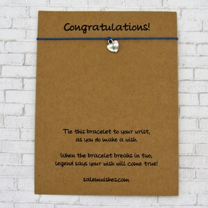 May include: A brown card with the text "Congratulations!" printed on it. The card has a blue string with a silver heart charm attached to it. The text on the card reads: "Tie this bracelet to your wrist, as you do make a wish. When the bracelet breaks in two, legend says your wish will come true! salemwishes.com"