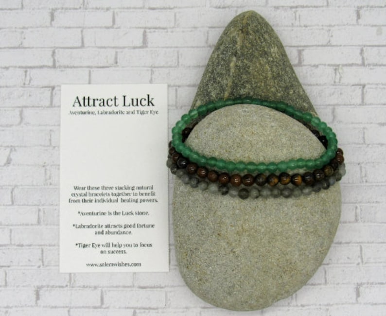 May include: Three stacking bracelets made of aventurine, labradorite, and tiger eye stones. The bracelets are on a gray rock with the text "Attract Luck" and a description of the stones' properties.