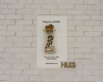 May include: A clear glass bottle with a cork stopper, labeled "Hug in a Bottle." The bottle contains small, colorful objects and the word "HUG" inside. A wooden cutout of the word "HUG" is below the card.