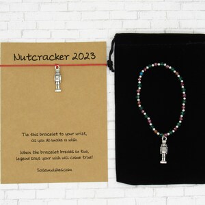 May include: A red string bracelet with a silver nutcracker charm. The bracelet comes in a black velvet pouch with a card that reads "Nutcracker 2023" and includes instructions on how to make a wish.