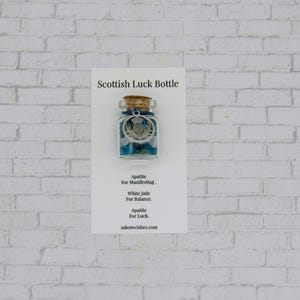 May include: A small clear glass bottle with a cork stopper, filled with blue apatite and white jade stones. A silver-toned Scottish thistle charm hangs from a blue string around the neck. The bottle is mounted on a white card labeled Scottish Luck Bottle.