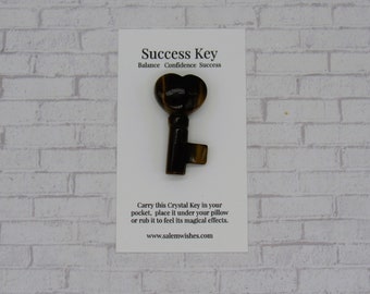 Può includere: Una chiave di cristallo di occhio di tigre marrone e dorata a forma di cuore. La chiave è su una carta bianca con il testo "Success Key" e "Balance Confidence Success". La carta dice anche "Carry this Crystal Key in your pocket, place it under your pillow or rub it to feel its magical effects. www.salemwishes.com"
