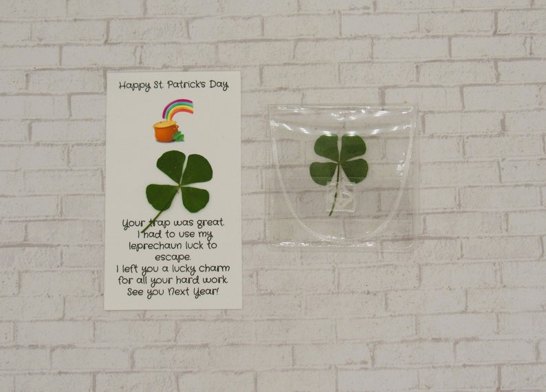 May include: A white card with the text "Happy St. Patricks Day" and a message about a leprechaun escaping a trap. The card is next to a clear plastic bag containing a four-leaf clover.