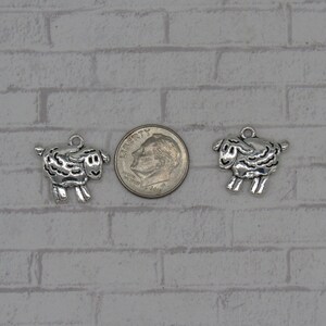 May include: Two silver-toned sheep charms, each with a detailed design, are shown next to a US dime for size comparison.