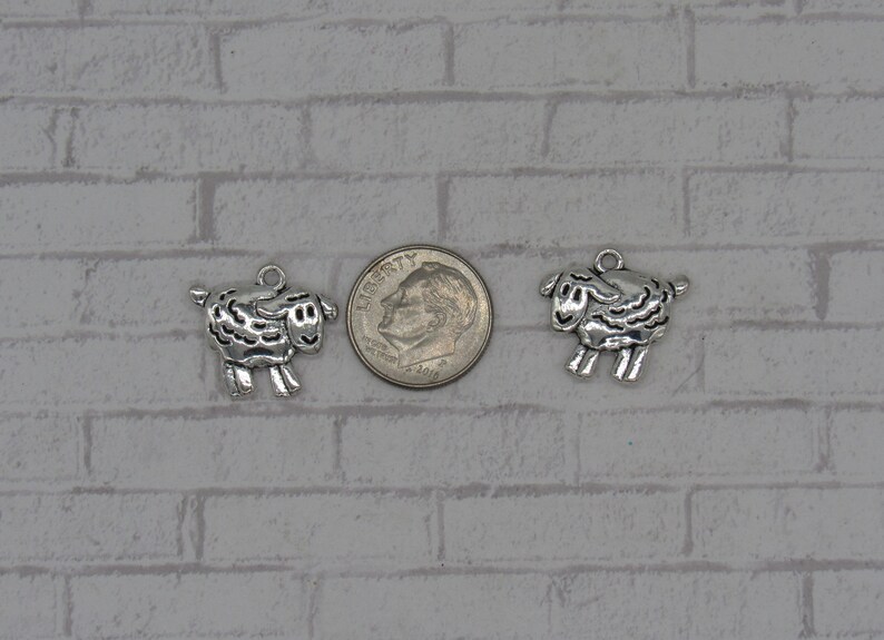 May include: Two silver-toned sheep charms, each with a detailed design, are shown next to a US dime for size comparison.