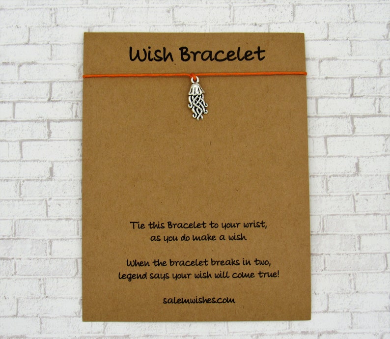 May include: A brown card with the text "Wish Bracelet" printed in black. The card has an orange string attached to it with a silver charm shaped like a jellyfish. The text on the card reads: "Tie this Bracelet to your wrist, as you do make a wish. When the bracelet breaks in two, legend says your wish will come true! salemwishes.com"