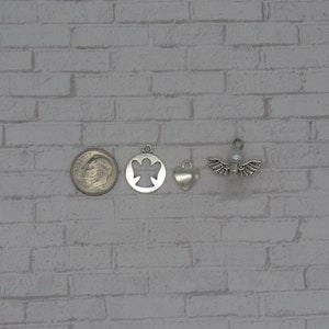 May include: Four silver charms, including an angel, a heart, and two angel wings with a pearl in the center. The charms are arranged in a row on a white brick background.