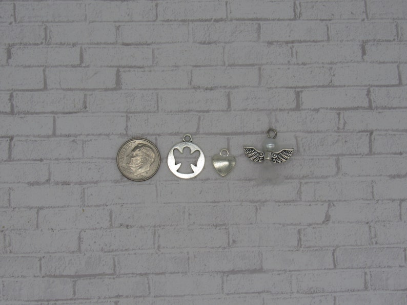May include: Four silver charms, including an angel, a heart, and two angel wings with a pearl in the center. The charms are arranged in a row on a white brick background.