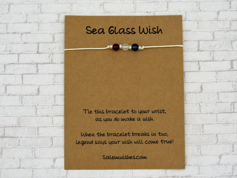 K&ouml;nnte beinhalten: Ein wei&szlig;es Armband aus Schnur mit roten, wei&szlig;en und blauen Glasperlen. Die Karte lautet "Sea Glass Wish" und enth&auml;lt Anweisungen, wie man mit dem Armband einen Wunsch macht. Die Karte enth&auml;lt auch die Website "Salemwishes.com".