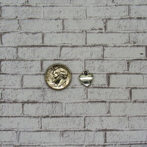 May include: A silver heart charm pendant, shown next to a US dime coin for size comparison. The charm has a loop at the top for attaching to a necklace or bracelet.