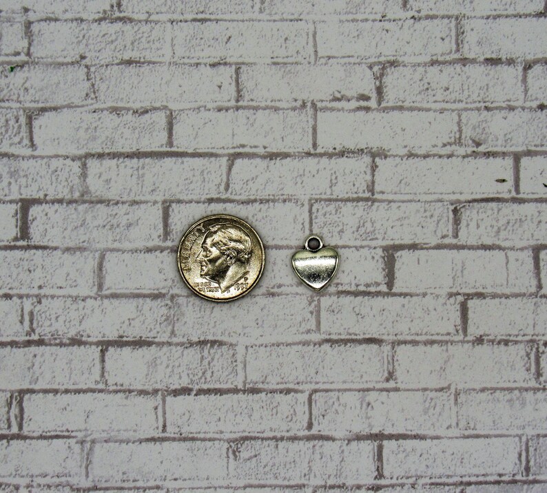 May include: A silver heart charm pendant, shown next to a US dime coin for size comparison. The charm has a loop at the top for attaching to a necklace or bracelet.