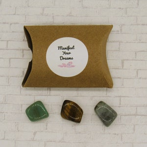 May include: Three tumbled stones in a brown paper pouch. The stones are green aventurine, tiger's eye, and grey labradorite. The pouch has a white sticker with the text "Manifest Your Dreams".