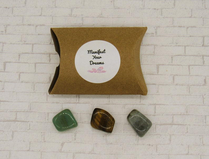 May include: Three tumbled stones in a brown paper pouch. The stones are green aventurine, tiger's eye, and grey labradorite. The pouch has a white sticker with the text "Manifest Your Dreams".